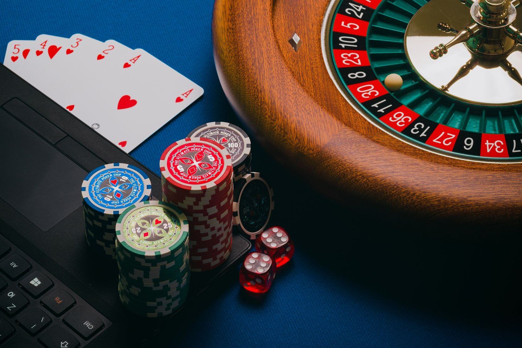Roulette wheel, casino chips, dice and playing cards next to a laptop keyboard